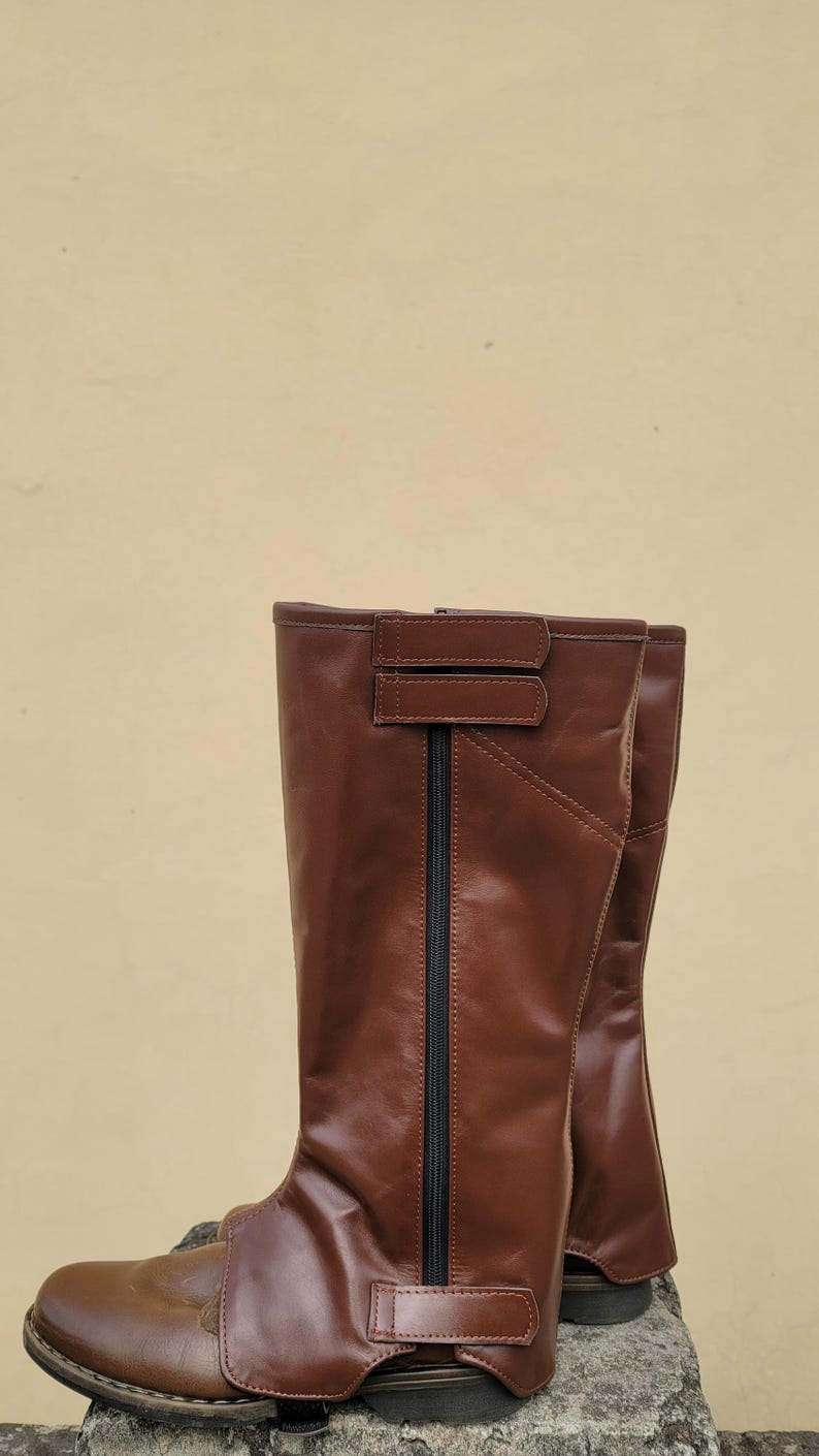 Puede incluir: Un par de botas de montar de cuero marr&oacute;n con una cremallera en la parte posterior y una correa en la parte superior.