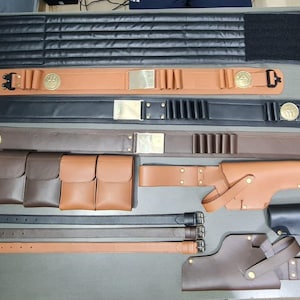May include: A collection of leather belts, pouches, and holsters in various shades of brown and black. Some belts feature decorative gold-colored accents. The items are arranged on a neutral surface, showcasing the craftsmanship of the leather goods.