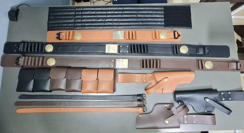 May include: A collection of leather belts, pouches, and holsters in various shades of brown and black. Some belts feature decorative gold-colored accents. The items are arranged on a neutral surface, showcasing the craftsmanship of the leather goods.
