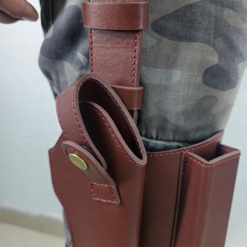 May include: A burgundy leather holster with a belt loop and a snap closure. The holster is attached to a matching leather belt. The stitching is a lighter shade of red. The holster is designed to be worn on the leg.