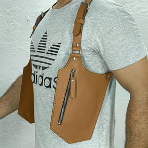 以下が含まれることがあります： 茶色の革製チェストホルスター。垂直のジッパーポケットが付いています。ホルスターには調節可能なストラップがあり、胸と肩に装着するように設計されています。ステッチが見え、革は滑らかな仕上がりです。