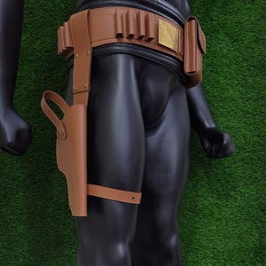 May include: A brown leather holster and belt set. The belt features multiple loops, a small pouch, and a decorative gold-colored buckle. The holster is attached to the thigh with a strap. The set is displayed on a black mannequin against a green background.
