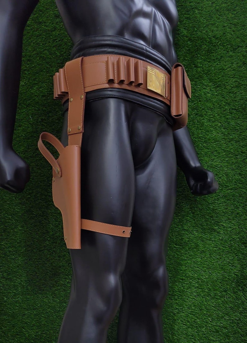 May include: A brown leather holster and belt set. The belt features multiple loops, a small pouch, and a decorative gold-colored buckle. The holster is attached to the thigh with a strap. The set is displayed on a black mannequin against a green background.