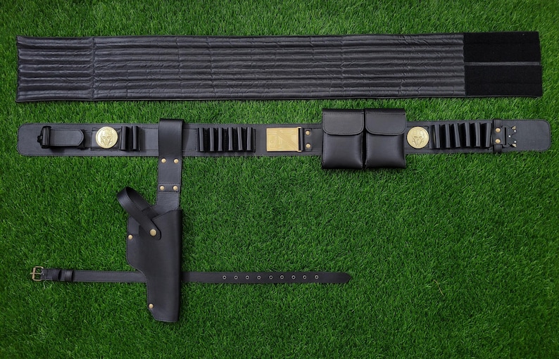 May include: A black leather utility belt with a holster, pouches, and cartridge holders. The belt features gold-colored accents and a separate padded back support. The items are displayed on a green surface.
