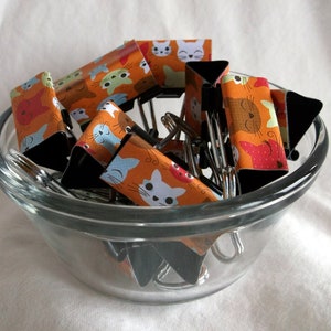 May include: A glass bowl filled with colorful binder clips featuring a cat design. The clips are orange, blue, and white with black accents.
