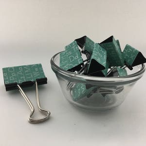 Puede incluir: Un bol de vidrio lleno de clips de carpeta verdes y negros. Los clips tienen letras blancas impresas en ellos. Un solo clip de carpeta plateado est&aacute; sobre la superficie blanca junto al bol.