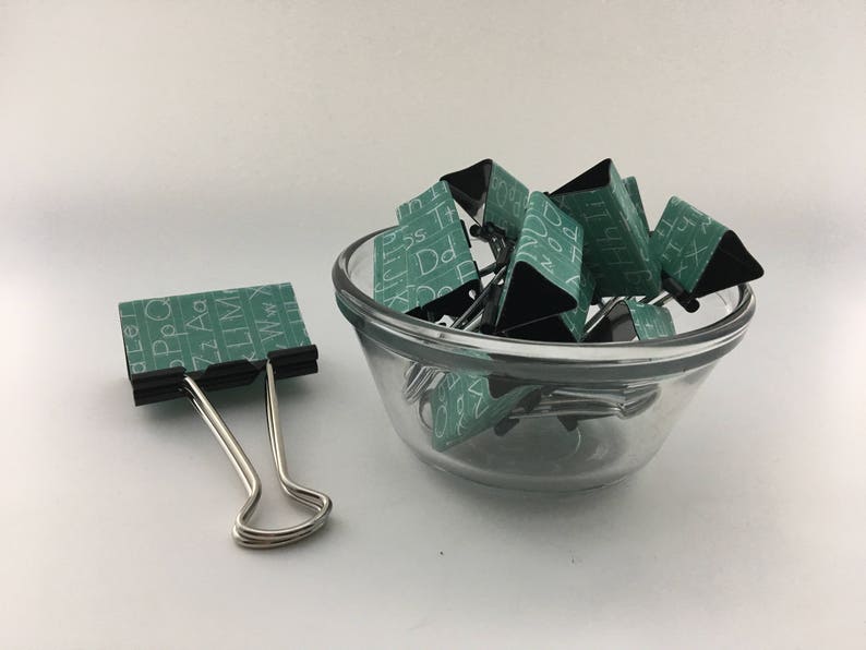 Puede incluir: Un bol de vidrio lleno de clips de carpeta verdes y negros. Los clips tienen letras blancas impresas en ellos. Un solo clip de carpeta plateado est&aacute; sobre la superficie blanca junto al bol.