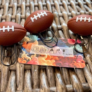 May include: Three brown football-shaped paper clips with white laces rest on a colorful card. The card has the word "CLIPART" and the text "by ferie". The clips are on a woven surface.