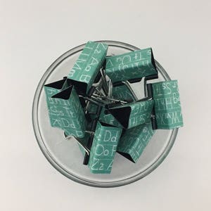 Puede incluir: Un bol de vidrio lleno de clips de carpeta verdes. Los clips est&aacute;n decorados con letras blancas, cada clip presenta una letra diferente del alfabeto.