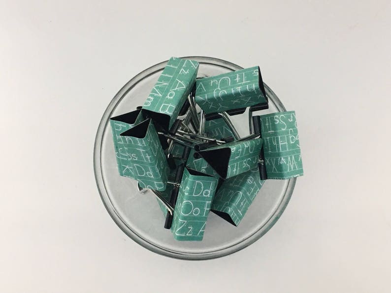 Puede incluir: Un bol de vidrio lleno de clips de carpeta verdes. Los clips est&aacute;n decorados con letras blancas, cada clip presenta una letra diferente del alfabeto.