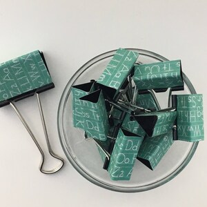 Puede incluir: Un bol lleno de clips de carpeta verdes con letras blancas en ellos. Las letras en los clips deletrean el alfabeto. Hay un solo clip de carpeta en el lado izquierdo de la imagen.