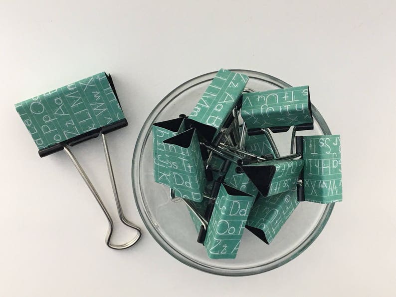 Puede incluir: Un bol lleno de clips de carpeta verdes con letras blancas en ellos. Las letras en los clips deletrean el alfabeto. Hay un solo clip de carpeta en el lado izquierdo de la imagen.