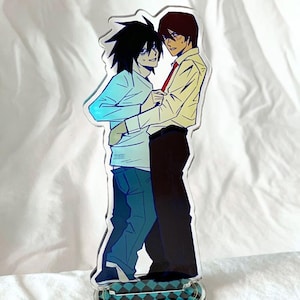 May include: Acrylic stand featuring two anime characters in a close embrace. One character has black hair and is wearing a light blue shirt and jeans. The other character has brown hair, a yellow shirt, and a red tie. The base has a blue and black checkered pattern.