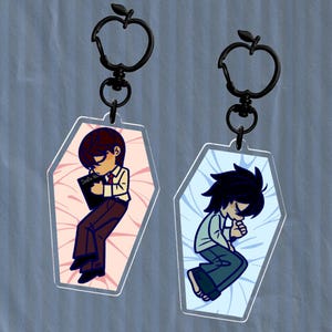 May include: Two acrylic keychains featuring anime characters in a coffin-shaped design. One keychain shows a character in a white shirt, brown pants, and a red tie, holding a book. The other keychain shows a character in a light green shirt and blue pants.