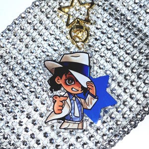 May include: A clear acrylic keychain featuring a cartoon illustration of a person wearing a white fedora, a white suit jacket, and a blue tie. The character is pointing forward. The keychain includes a gold-colored star-shaped clasp and a blue star charm.