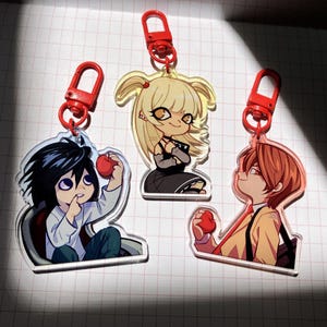 May include: Three acrylic keychains featuring anime characters. Each keychain has a red clasp and depicts a different character holding a red apple. The characters have distinct hairstyles and outfits, with varying color palettes.