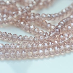 Puede incluir: Primer plano de hebras de cuentas facetadas de color púrpura claro. Las pequeñas cuentas redondas tienen un aspecto cristalino. Están ensartadas, creando un efecto fluido y texturizado. Ideal para la creación de joyas.