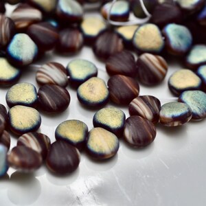 May include: A collection of flat, oval-shaped beads in shades of brown, cream, and blue. The beads have a striped pattern and a slightly iridescent finish. They are scattered on a white surface, suggesting they are for jewelry making or crafts.
