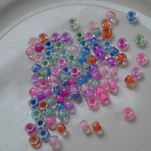 Op de afbeelding: Een verzameling kleurrijke, doorschijnende plastic kralen in verschillende tinten roze, blauw, groen en oranje. De kralen zijn verspreid over een wit oppervlak.