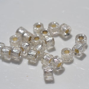 May include: A group of silver and clear glass beads with a metallic finish. The beads are round and have a hole in the centre. They are scattered on a white surface.