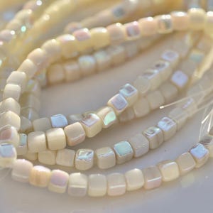 May include: A strand of small, square-shaped beads in a creamy off-white color. Each bead features a smooth, rounded finish with an iridescent, pearlescent inlay on one side that reflects soft hints of blue, pink, and purple light in the sun.