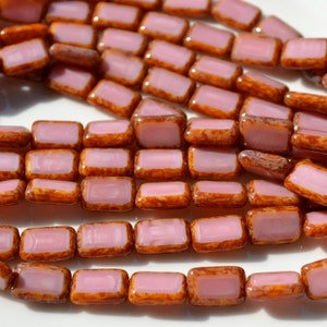 May include: Close-up of rectangular beads with a light pink center and a brown, textured border. The beads are strung together, showcasing their shape and color variations. These beads are ideal for jewelry making.