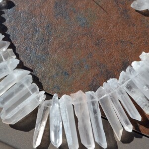 May include: A strand of clear quartz crystal points, arranged in a semi-circle on a brown textured surface. The points are various lengths and have a natural, raw appearance.