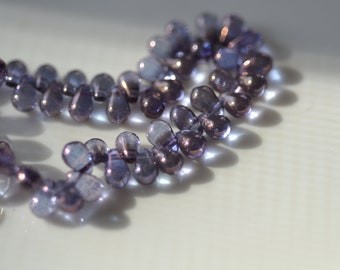 May include: A strand of iridescent, teardrop-shaped beads in shades of purple and blue. The beads are strung together, creating a delicate and elegant appearance. The beads are approximately 0.5 inches in length, and are displayed on a white surface.