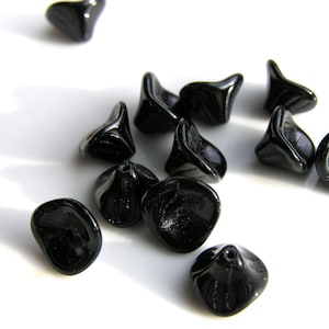 Puede incluir: Un conjunto de cuentas de vidrio negro brillante. Las cuentas tienen una forma org&aacute;nica &uacute;nica con una base redondeada y una parte superior acampanada. Est&aacute;n dispersas sobre una superficie blanca, con la luz reflej&aacute;ndose en sus superficies lisas.