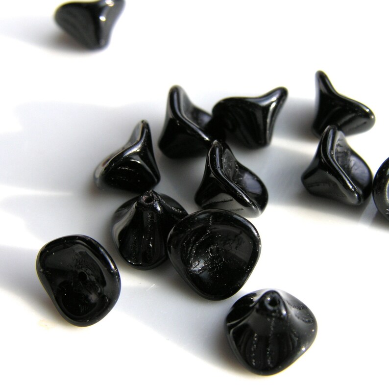 Puede incluir: Un conjunto de cuentas de vidrio negro brillante. Las cuentas tienen una forma org&aacute;nica &uacute;nica con una base redondeada y una parte superior acampanada. Est&aacute;n dispersas sobre una superficie blanca, con la luz reflej&aacute;ndose en sus superficies lisas.