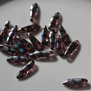 May include: A collection of teardrop-shaped glass beads in shades of purple and iridescent blue. Each bead features a series of colorful, circular accents. The beads are scattered on a white surface, creating a visually appealing arrangement for jewelry making.