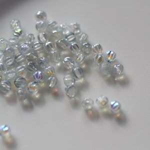 May include: A pile of iridescent clear glass beads with a faceted design. The beads are scattered on a white surface.