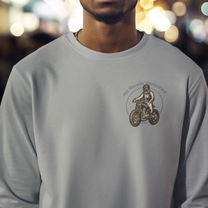 以下が含まれることがあります： クルーネックのライトグレーのスウェットシャツ。スウェットシャツの左胸上部には、「No Roads Required」の文字とバイクに乗った人のイラストが描かれた小さな円形のグラフィックがあります。画像に写っている人物はグレーのスウェットシャツを着ています。