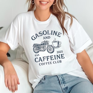 以下が含まれることがあります： 白のTシャツ。ダークブルーのグラフィックデザインで、オートバイ、コーヒーカップ、そして「GASOLINE AND CAFFEINE COFFEE CLUB」の文字と「2025」の年号が描かれています。カジュアルなスタイルです。