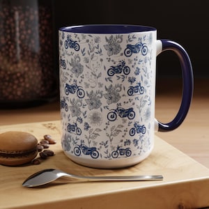 May include: White ceramic mug with a blue interior and handle. The mug features a pattern of blue motorcycles and floral designs. A spoon and a macaron are on a wooden surface next to the mug.