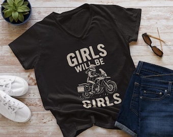 May include: Black v-neck t-shirt with the text "GIRLS WILL BE GIRLS" and a motorcycle graphic. The shirt is paired with blue denim shorts, white sneakers, and sunglasses on a wooden surface.