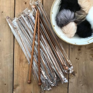 Può includere: Un insieme di ferri da maglia in bambù in confezioni di plastica trasparente, accanto a una ciotola decorativa piena di pompon di finta pelliccia. Gli aghi sono di varie lunghezze e hanno estremità arrotondate.