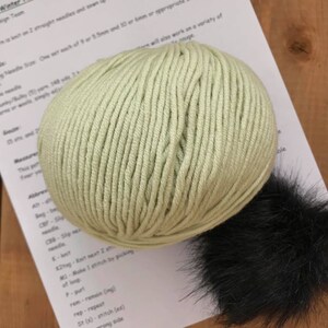 May include: A ball of light green yarn with a black faux fur pom pom. The yarn is for knitting or crocheting. The yarn is next to a piece of paper with knitting instructions.