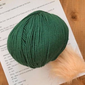 May include: A ball of dark green yarn and a small, fluffy, peach-coloured pom-pom. The yarn is for knitting a hat. The instructions for the hat are visible in the background.