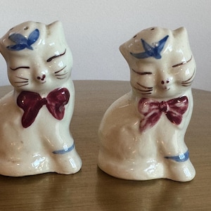 May include: A pair of vintage ceramic cat salt and pepper shakers. The cats are cream-colored with closed eyes, a burgundy bow tie, and a blue accent on their heads and feet. The shakers are designed to look like seated cats.