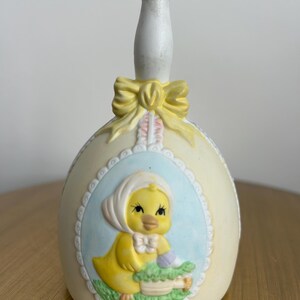 May include: A ceramic bell with a cream-colored egg shape, featuring a yellow chick in a blue oval frame. The bell has a white handle and a yellow bow. The chick is wearing a white bonnet and holding a basket.