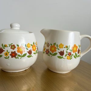 May include: A cream-colored ceramic sugar bowl with a lid and a matching creamer pitcher. Both pieces feature a repeating horizontal band of orange, yellow, and brown checkered flowers with green stems and leaves encircling the midsection.