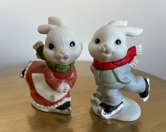 May include: Two ceramic bunny figurines. One bunny wears a red and white dress with a green scarf, the other wears a gray suit and red scarf. Both bunnies are ice skating and have black eyes.