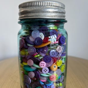 May include: A clear glass jar filled with a colorful assortment of buttons. The buttons are various sizes and shades of purple, pink, yellow, and green. The jar has a silver metal lid and sits on a gold surface.