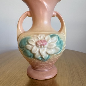 May include: A pink ceramic vase with two handles and a white flower design. The vase has a textured surface and a wide mouth.