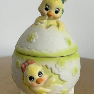 May include: A ceramic Easter egg-shaped container with a lid, featuring a yellow chick emerging from the egg. The chick wears a chef's hat and has a red bow. The container is white with green accents and floral details.