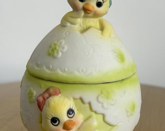 May include: A ceramic Easter egg-shaped container with a lid, featuring a yellow chick emerging from the egg. The chick wears a chef's hat and has a red bow. The container is white with green accents and floral details.