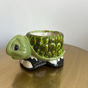 May include: A ceramic turtle-shaped planter with a glossy light green head and a textured, mottled green and brown shell. The base of the planter is black with white accents, and the interior is a smooth, off-white glazed finish.
