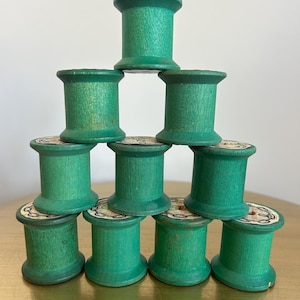 May include: A pyramid stack of ten cylindrical wooden spools painted in a vibrant green color. Each spool features a flat top and bottom with a visible wood grain texture. The spools are arranged in four rows, with circular paper labels on the ends.