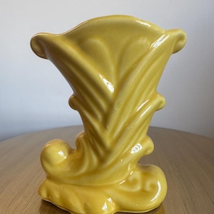 May include: A yellow ceramic vase with a unique, stylized design. The vase features a flared top, a central column with vertical ridges, and a decorative base with swirling details. The glossy finish enhances the vase's aesthetic appeal.